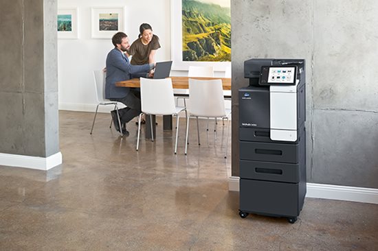 So finden Sie den richtigen Drucker fürs Büro: Offenes Büro mit einem Multifunktionsdrucker, Mann und Frau sitzen am Besprechungstisch und arbeiten gemeinsam